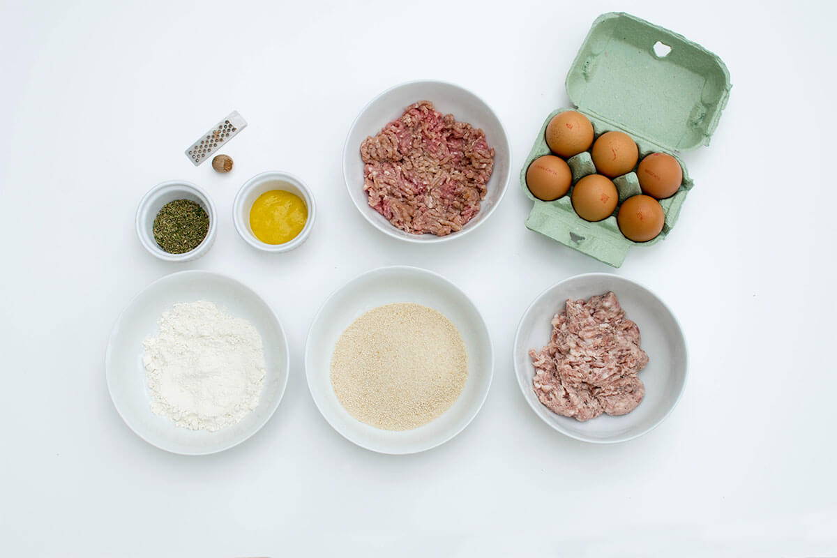 A crate of 6 eggs next to a bowl of sausage meat, a bowl of turkey mince, a bowl of flour, a bowl of breadcrumbs, dried herbs, nutmeg and mustard