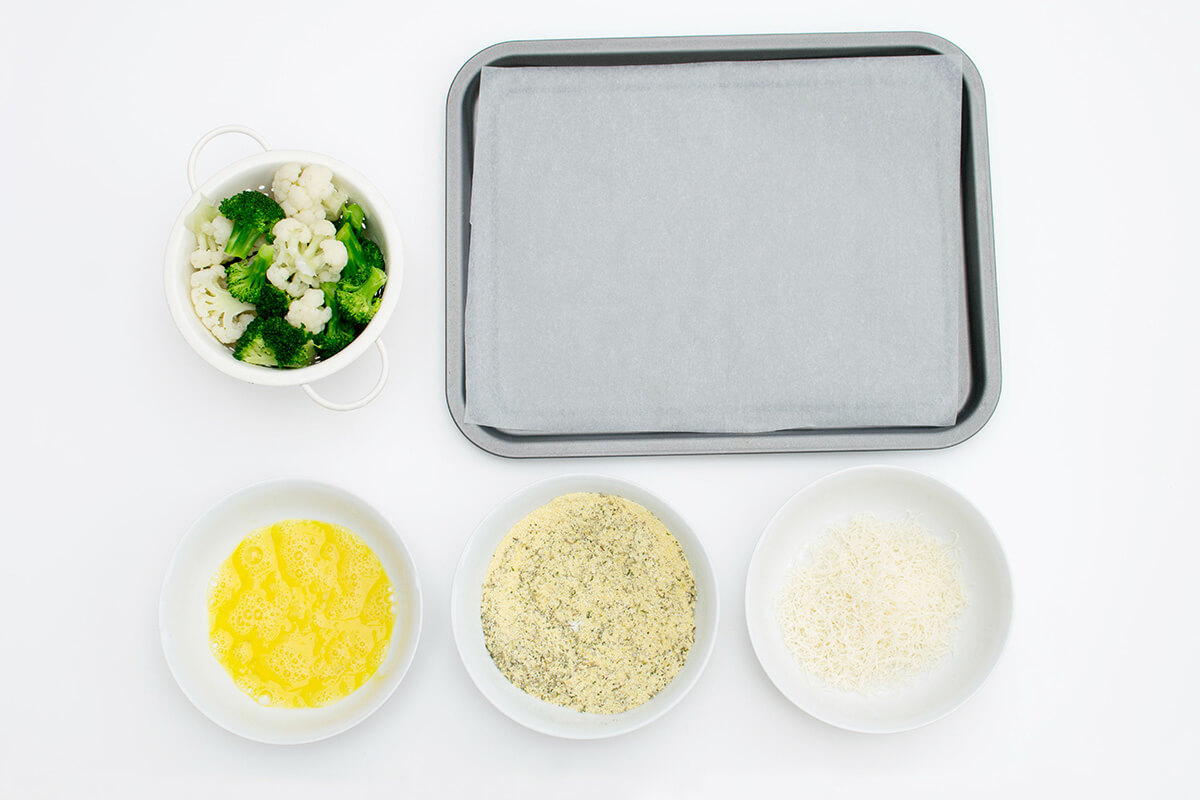 An oven tray next bowl of cauliflower and broccoli florets, a bowl of beaten egg, a bowl of polenta and herb mix and a bowl of parmesan