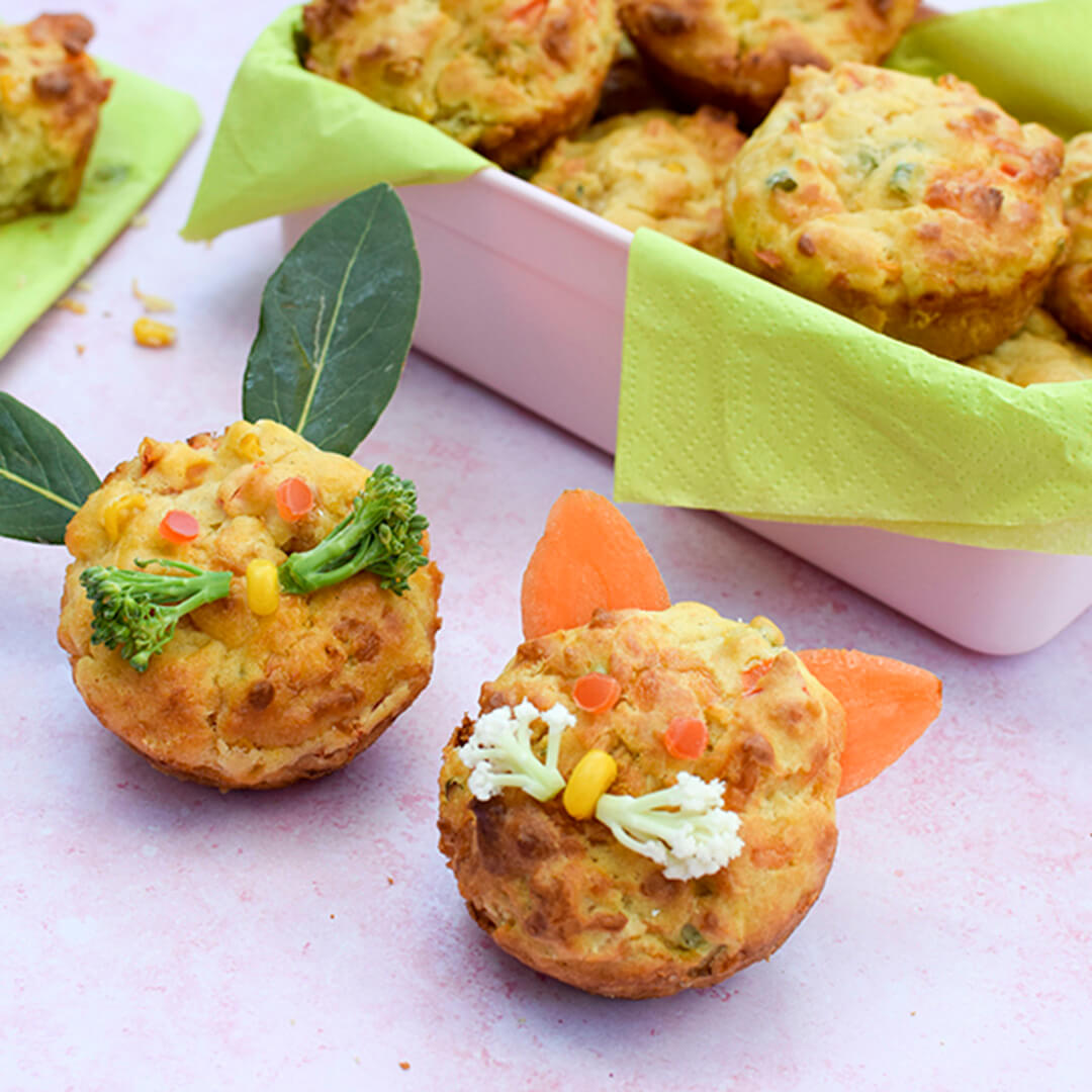 A container of Savoury Vegetable Muffins for Babies &amp; Toddlers next to two savoury vegetable muffins - one of them has a sweetcorn nose with small broccoli floret whiskers and herb leaves for ears, the other has a sweetcorn nose, small cauliflower floret whiskers and carrot slices for ears